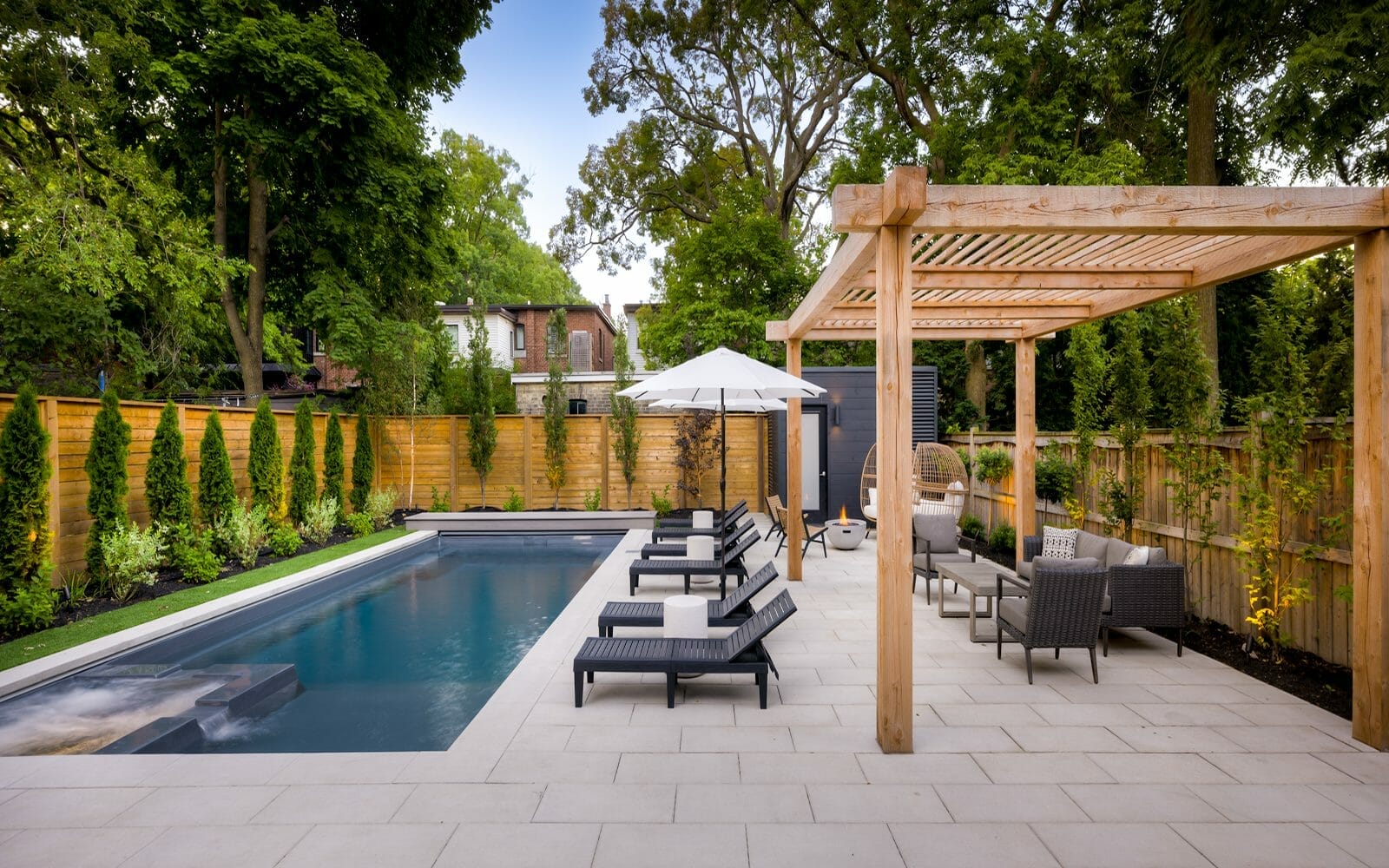 Complete Modern Backyard Landscape Design Project, Featuring, Stone Interlocking, Woodworking Pergola, Fiberglass Pool with Swim Spa, Outdoor Fireplace, Elegant Outdoor Furniture Alongside the Pool, Grey Storage Shed Near the Back of the Property, Privacy Fence, and Small Trees and Shrubs.