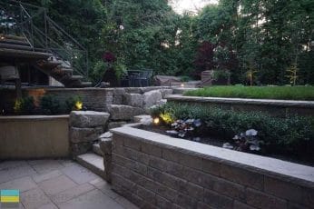 M.E. Contracting built stone steps out of a basement walkway, as well as steps leading to an unseen deck, Also shown is landscaping with lighting.